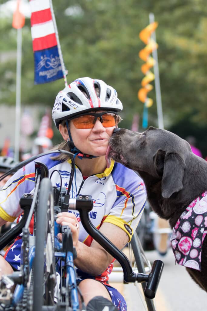 Holly, wearing a cycling uniform, celebrates the end of a race with a kiss on the check from her service dog. 