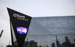 Exterior view of U.S. Bank Stadium with large glass windows and angular black facade, displaying a video screen showing logos for the Green Bay Packers and Minnesota Vikings.
