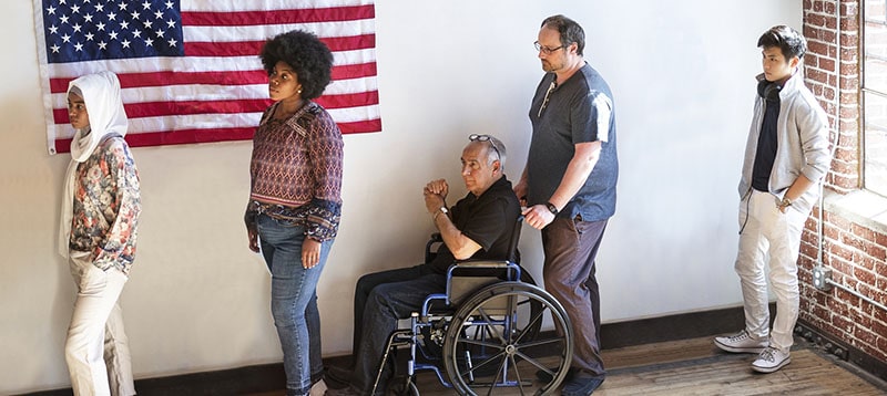 People wait in line to vote with an American flag behind them. In the middle is a man in a wheelchair being pushed by another man.