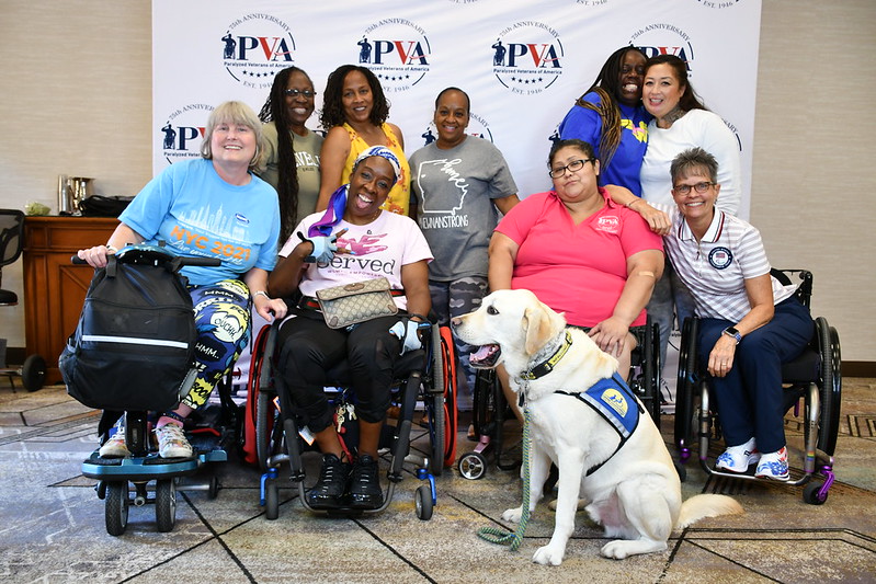 Women veterans in wheelchairs and a service dog pose in front of a PVA banner at the 2024 Women Veterans Empowerment Retreat