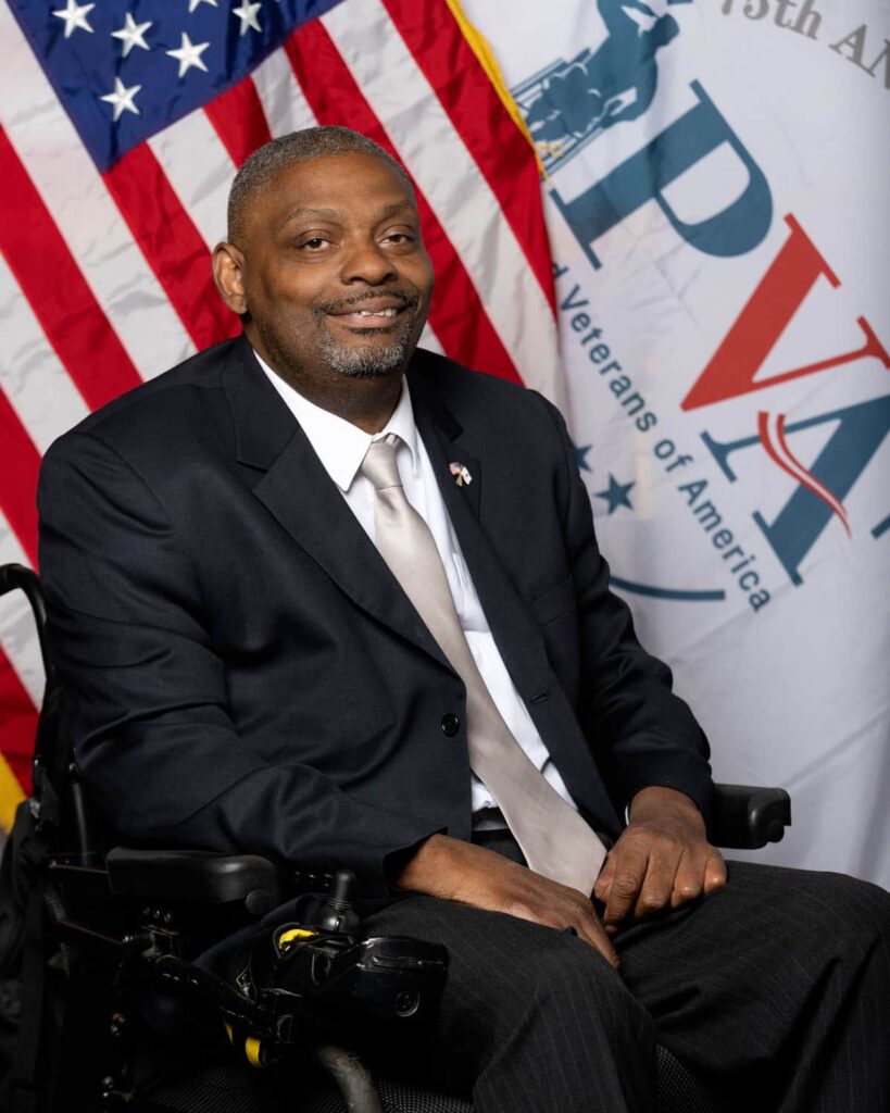 Robert Thomas' official PVA portrait shows him in his power chair, wearing a suit, in front of an American flag and a PVA flag.