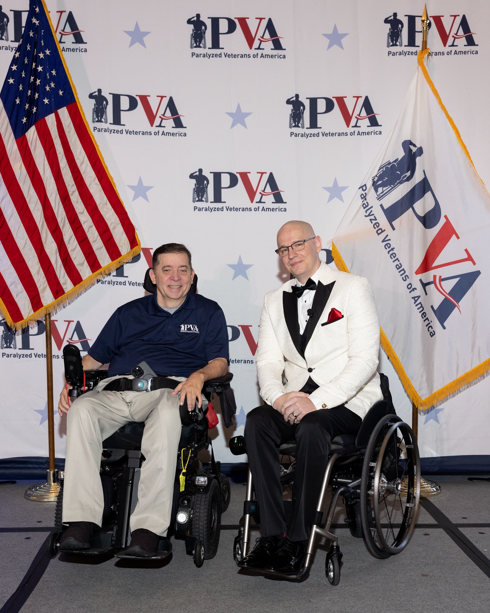 Immediate Past President Charles Brown and Chief Operating Officer Shaun Castle pose in a power wheelchair and manual chair with PVA in the background and an American flag and flag with a PVA logo on either side of them