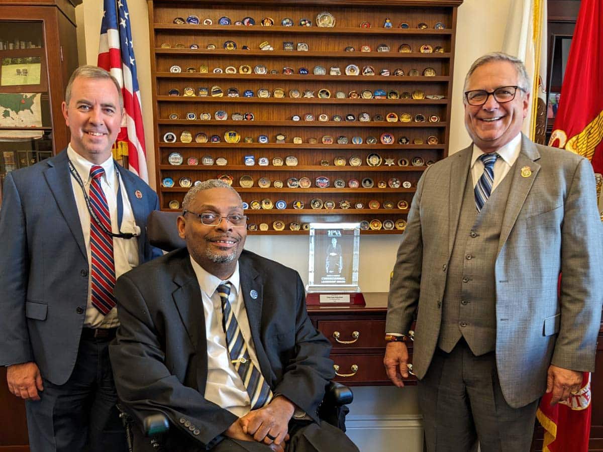From left to right: PVA Chief Executive Officer Carl Blake, PVA National President and Chairman of the Board Robert Thomas, in a power wheelchair, and Congressman Mike Bost smile at the camera. On the table behind the trio, below a shelving unit that holds various Challenge Coins, is the illuminated glass Gordon H. Mansfield Congressional Leadership Award.