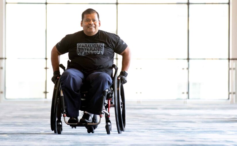 Army Veteran Michael wheels his wheelchair down a hall in front of a wall of windows while smiling