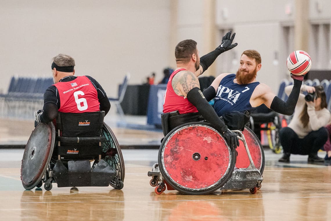 One man in a wheelchair throws a ball while another wheelchair user blocks him in a gymnasium