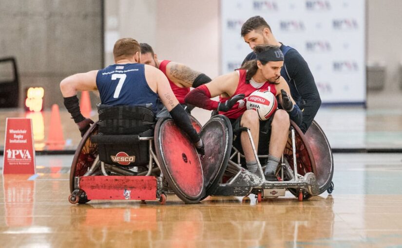 A man in a wheelchair wearing a red rugby holds a rugby ball surrounded by other wheelchair rugby players in a gymnasium