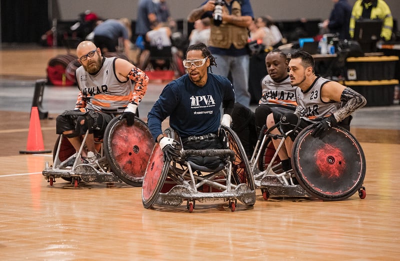 Four wheelchair rugby players look intently to the side as the ball passes by.