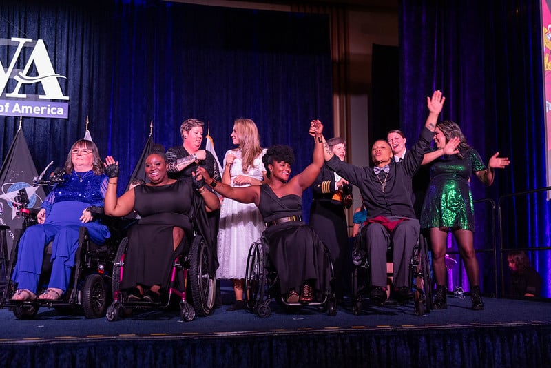 Women Veterans hold hands and sing on Stage at the PVA Igniting Change Gala. They are dressed in formal attire and smiling with their arms raised.