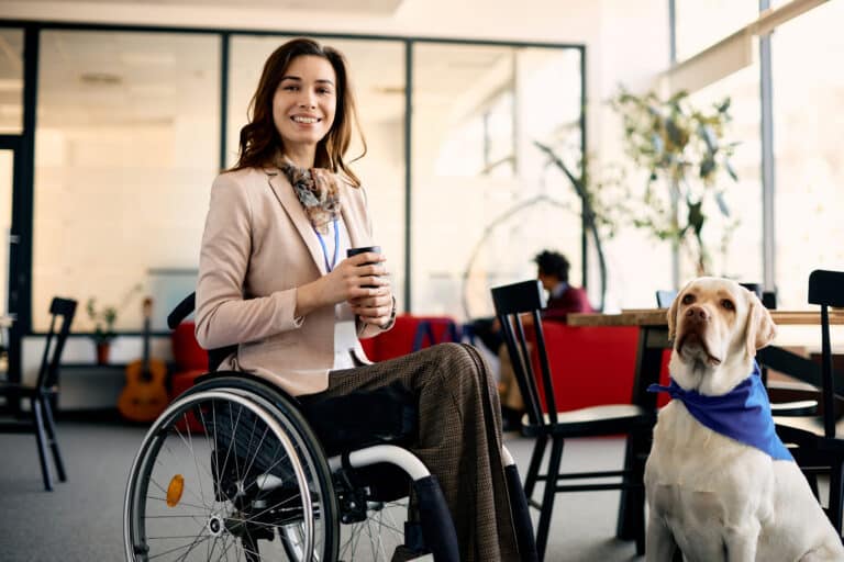 A professional woman in a manual wheelchair poses in the office, with her service dog next to her.