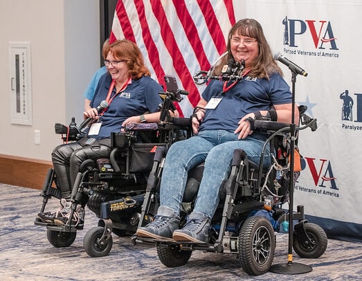 PVA leaders Tammy Jones and Anne Robinson, both in power wheelchairs, smile during a presentation at the women's retreat. There is a PVA logo wall and an American flag behind them.
