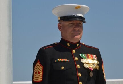 A man stands tall wearing a black military dress uniform with several metals on the jacket. he is also wearing a white military dress hat.