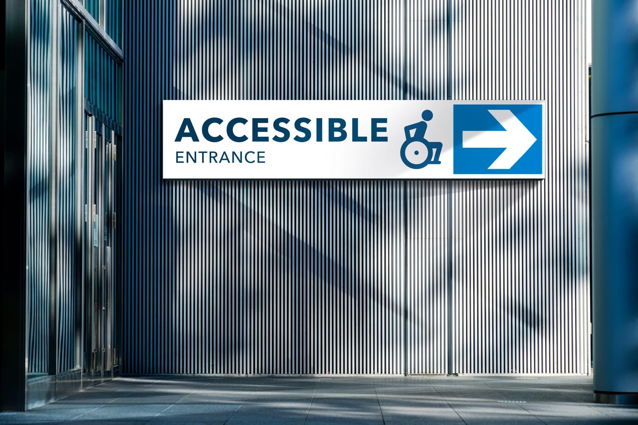 A wall-mounted sign with a blue and white design points to the accessible entrance. The sign features an arrow pointing right and a wheelchair icon above the text "ACCESSIBLE ENTRANCE." The background consists of vertical corrugated metal panels.