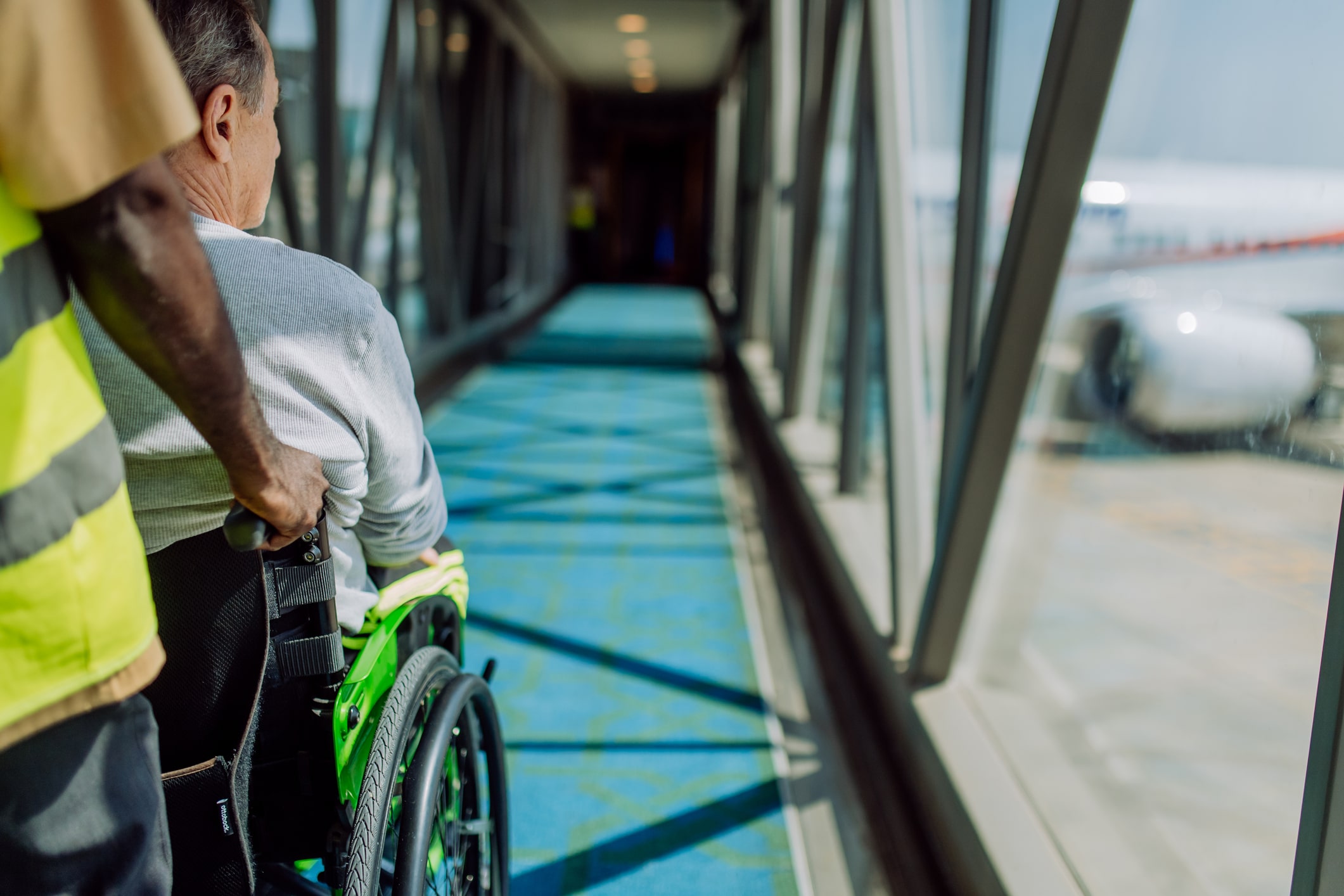 A person in a wheelchair is being assisted by an airport staff member along a glass-walled corridor leading to an airplane. The corridor has blue flooring with sunlight streaming through the windows creating patterns. An airplane is visible through the windows.