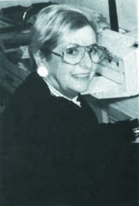A woman with short, light-colored hair and large glasses smiles at the camera while seated at a desk. She is wearing a dark jacket and light-colored blouse. In the background, there appears to be office equipment, including a typewriter or printer.