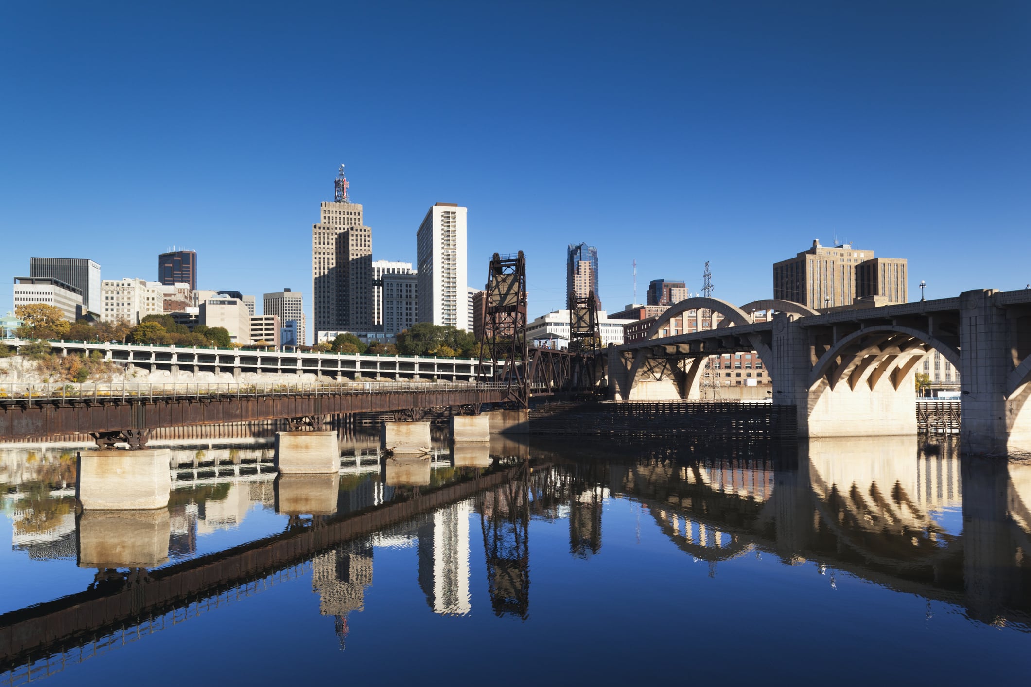 A cityscape featuring a railway bridge and high-rise buildings reflected in the calm water of a river beneath a clear blue sky. The scene showcases a combination of modern and historical architectural styles.