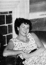 A woman with short, dark hair is sitting and smiling at the camera. She is wearing a patterned sleeveless dress and appears relaxed, leaning back slightly. The background shows a fireplace and part of a window. The image is in black and white.