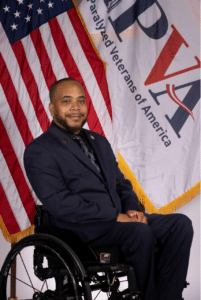 A man in a navy blue suit sits in a wheelchair in front of an American flag and a banner that reads "Paralyzed Veterans of America." He is smiling and has a short beard.