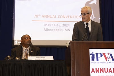 A man in a suit stands at a podium labeled "PVA" with a microphone, speaking at the 78th Annual Convention. Another man, seated at a table, looks on. The background shows a screen with text indicating the event dates and location: May 14-18, 2024, Minneapolis, MN.