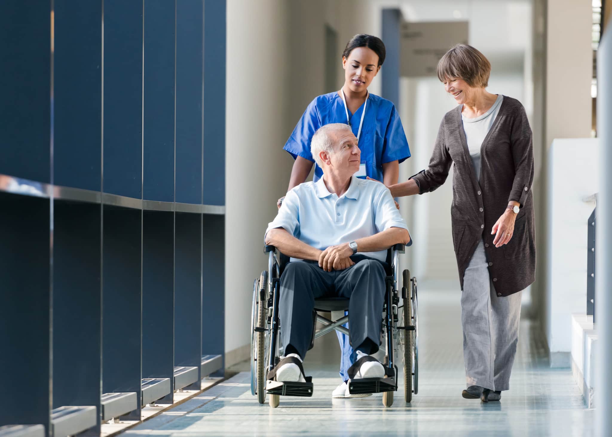 A nurse in blue scrubs pushes an elderly man in a wheelchair down a corridor as an older woman walks beside them, smiling and touching his shoulder. The setting appears to be a modern healthcare facility.