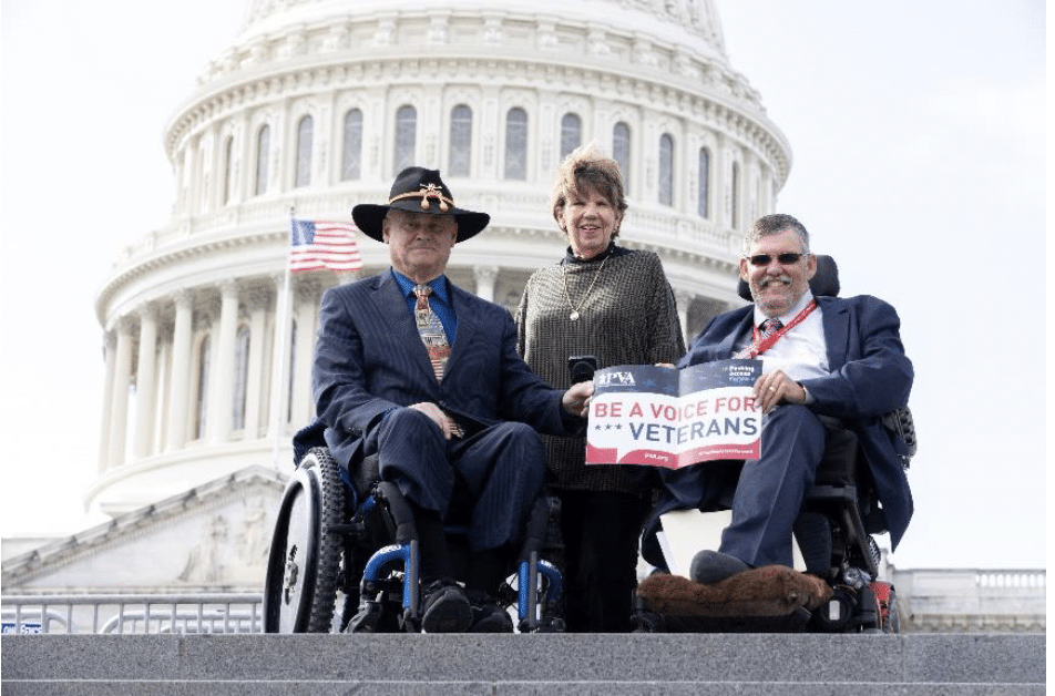 Three individuals, two seated in wheelchairs and one standing behind them, pose in front of the U.S. Capitol. One person holds a sign that reads "BE A VOICE FOR VETERANS." An American flag is visible on the left.
