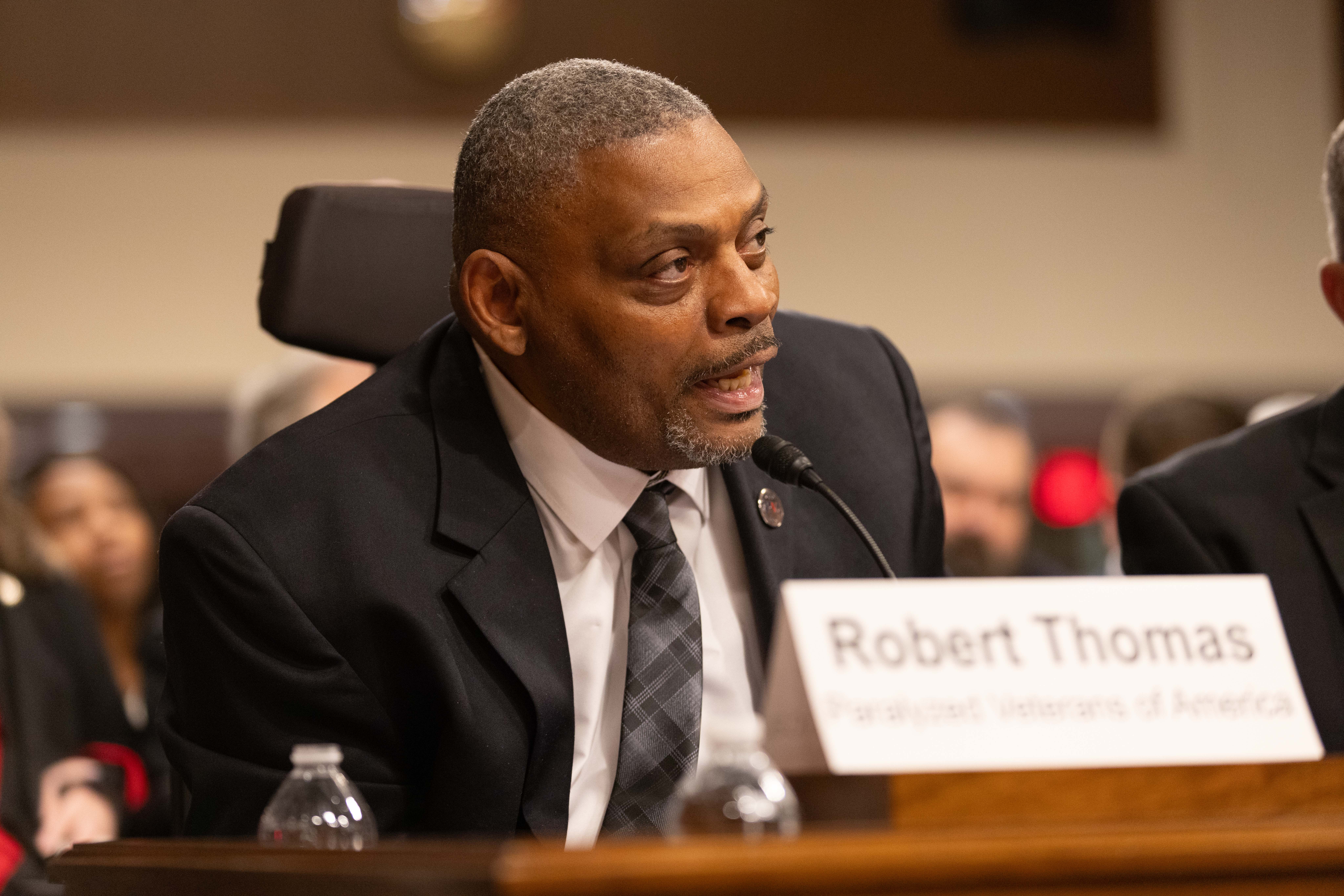 A man in a suit speaks into a microphone at a formal event or meeting. He wears a black tie and is seated at a panel. The nameplate in front of him reads "Robert Thomas" and "Paralyzed Veterans of America.