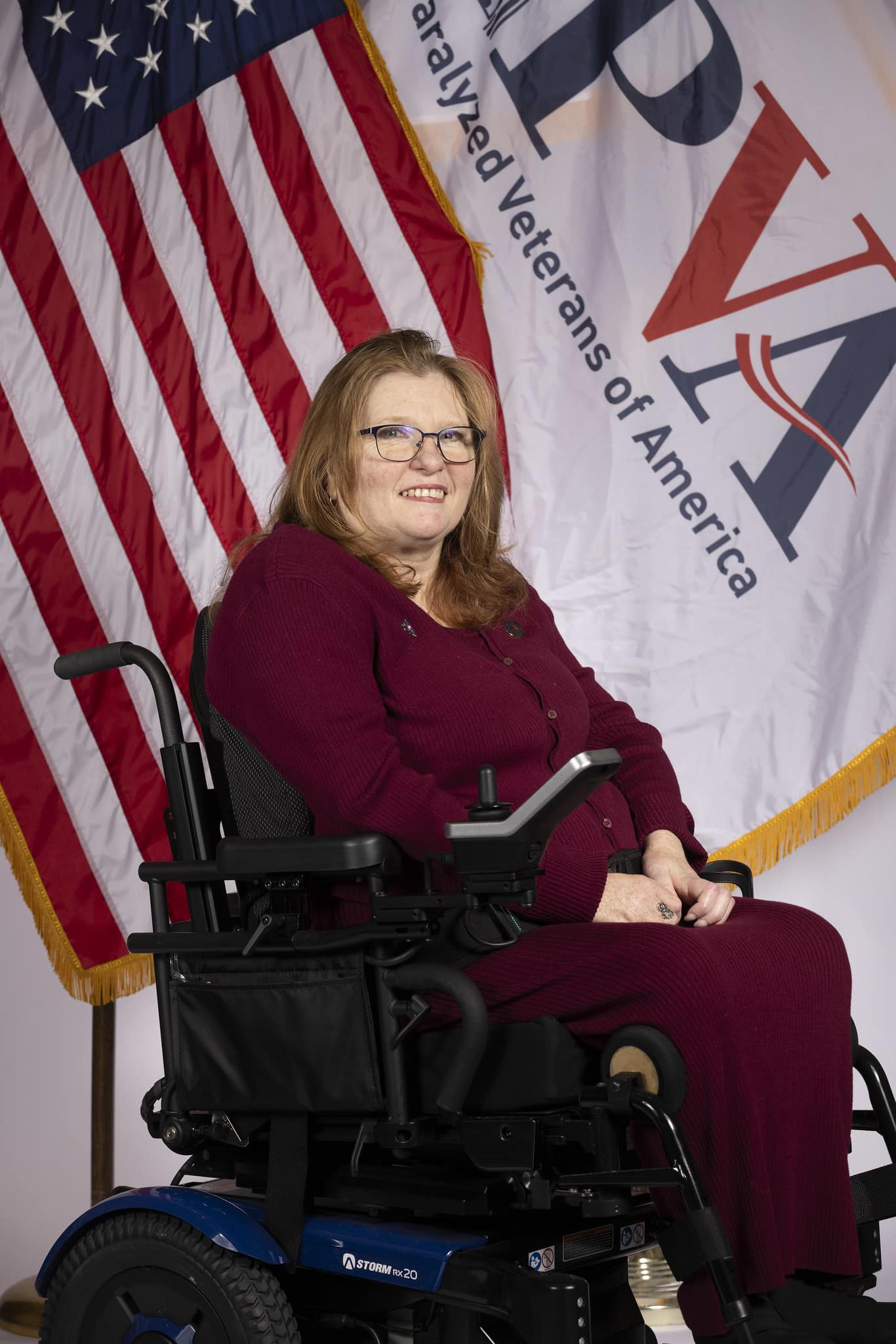 A woman with long, light brown hair and glasses is sitting in a wheelchair. She is wearing a maroon outfit and smiling. Behind her is an American flag and a large banner with the text "Paralyzed Veterans of America".