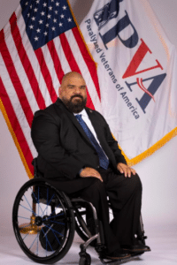 A man with a beard in a suit and tie sits in a wheelchair in front of an American flag and a banner with the logo of the Paralyzed Veterans of America (PVA).