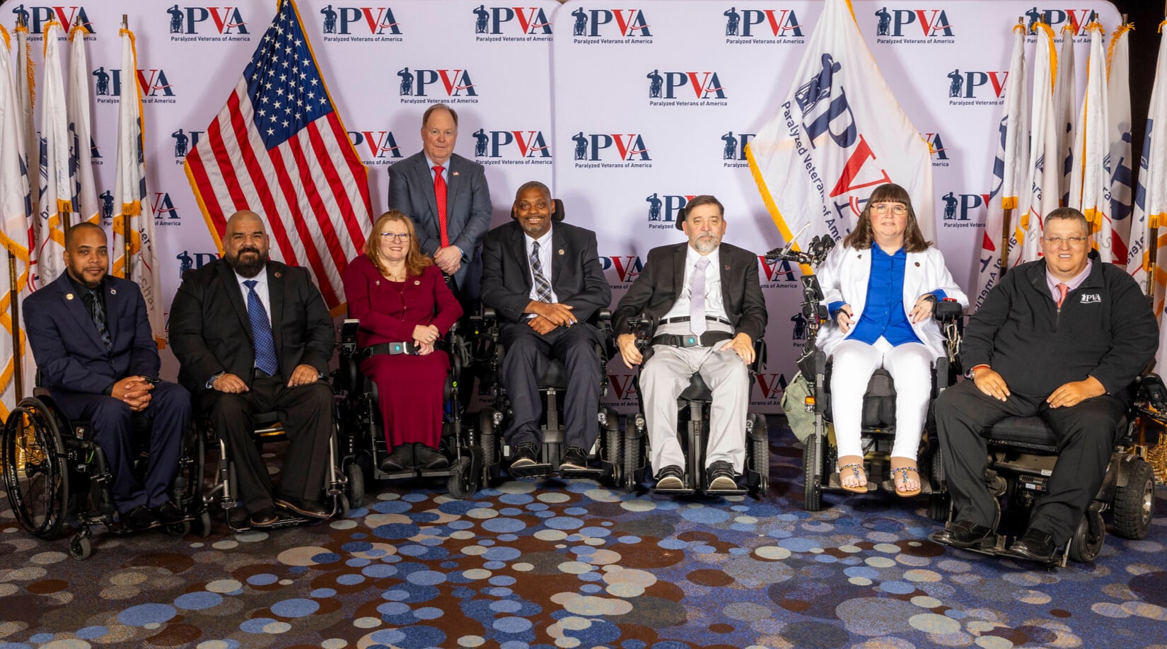 A group of eight individuals, some in wheelchairs, pose for a photo in front of a backdrop with PVA logos and flags. They are dressed in formal attire and appear to be at an event or conference.