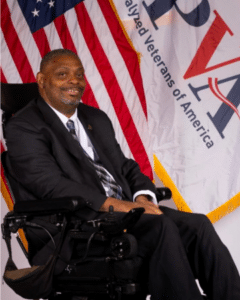 A man with short hair and a beard, dressed in a dark suit and plaid tie, is seated in a motorized wheelchair. He is smiling and positioned in front of a backdrop that features the American flag and a banner with the text "Paralyzed Veterans of America.