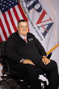 A man sits in a wheelchair, smiling, wearing glasses, a black sweater with the PVAV logo, and a striped tie. Behind him are a U.S. flag and the Paralyzed Veterans of America flag.