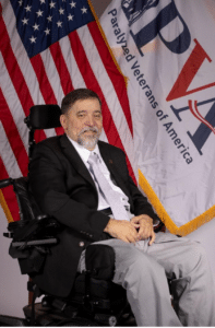 A man with a beard and mustache is seated in a motorized wheelchair, wearing a black blazer, white shirt, and gray pants. Behind him, there is an American flag and a "Paralyzed Veterans of America" banner. He is smiling softly.