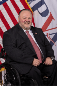 A smiling man with a beard, wearing a dark suit and red tie, sitting in a wheelchair. He is in front of an American flag and a backdrop with large, partially visible letters.