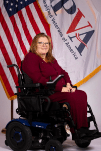 A woman with long hair and glasses is seated in a motorized wheelchair. She is wearing a burgundy outfit and smiling at the camera. Behind her are the American flag and a Paralyzed Veterans of America banner.