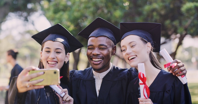 Three graduates wearing black caps and gowns smile while taking a selfie outdoors. They each hold rolled-up diplomas tied with red ribbons, celebrating their accomplishment. Trees and greenery are visible in the background.