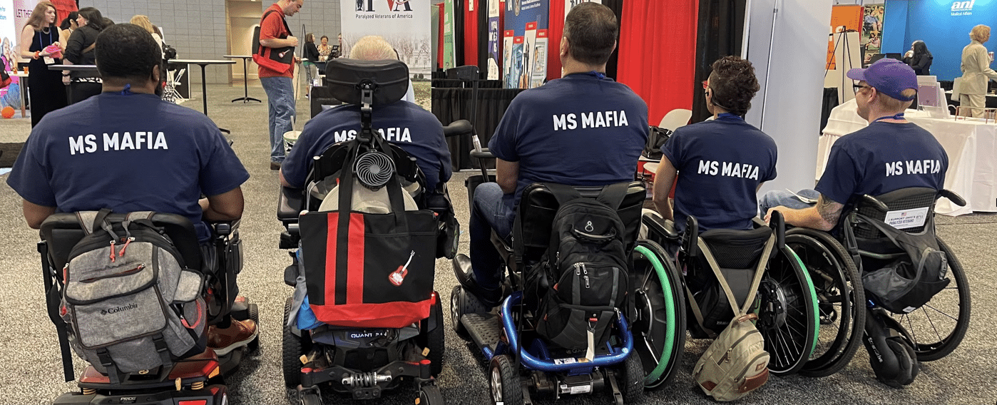 A group of five people in wheelchairs wearing matching navy blue T-shirts with "MS MAFIA" printed on the back. The scene appears to be at an indoor convention or event space, with various booths and displays visible in the background.