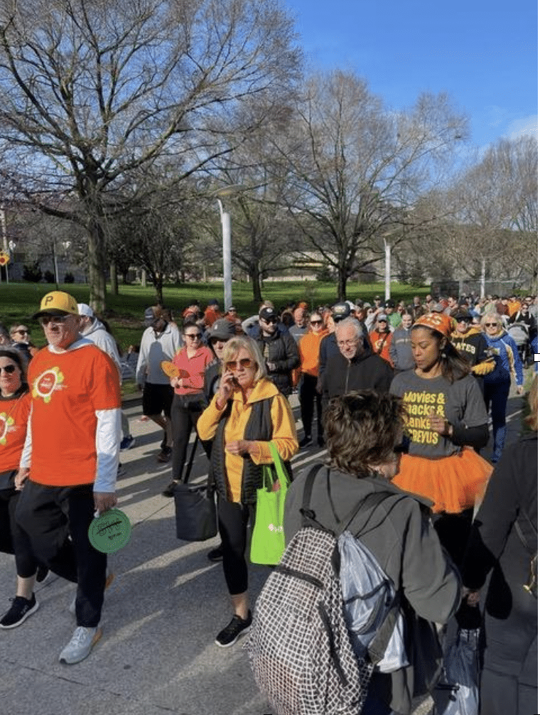 A large group of people, some wearing orange shirts, walk on a path in a park on a sunny day. The participants appear to be part of an organized event. A few are holding discs or wearing backpacks, and some trees in the background are bare.
