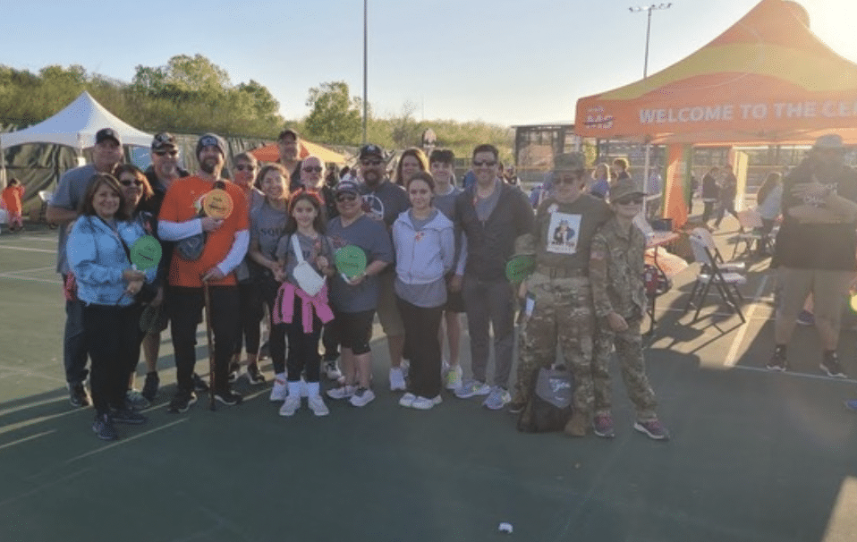 A group of people stands together outdoors on a sports court, many holding pickleball paddles. They are dressed in casual clothes, and some wear event T-shirts. Behind them is a tent and an orange canopy with the words "WELCOME TO THE CEL" partially visible.