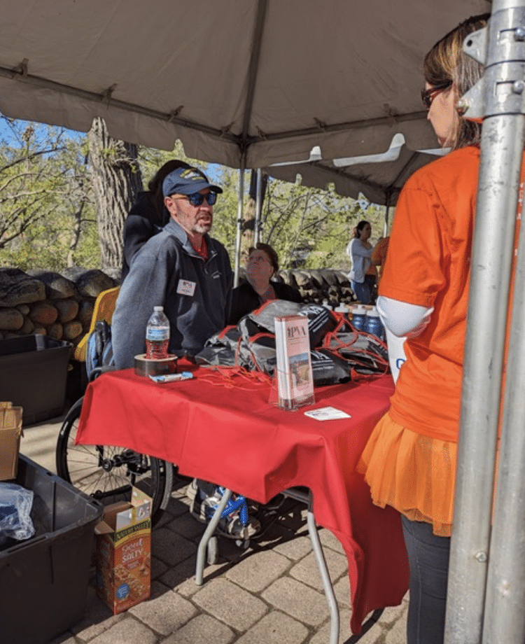A man in a wheelchair and a woman sit under a canopy at a red table with items and informational brochures on it. A woman in an orange shirt and tutu stands in front of the table. The setting appears to be outdoors on a sunny day.