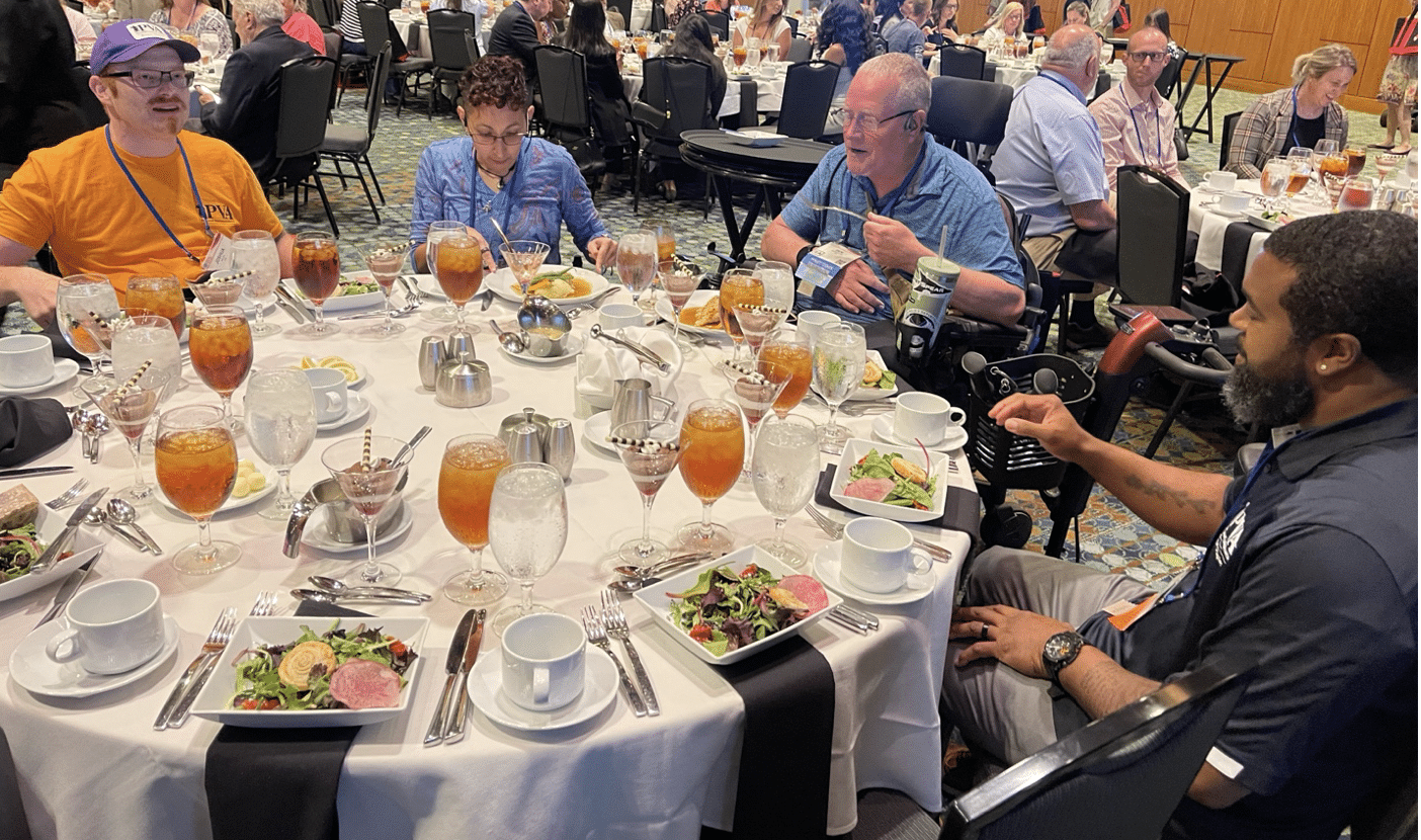 A group of four people sitting around a round table set with salads, glasses of iced tea, and various dining utensils. The group appears to be engaged in conversation while other dining tables and attendees are visible in the background.
