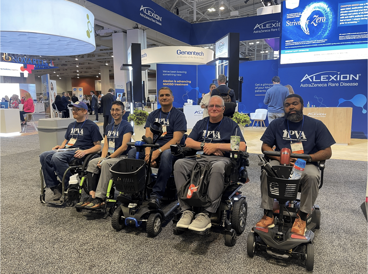 Five individuals using wheelchairs pose for a group photo at a conference booth area. They wear matching navy blue shirts with "PVA" printed on them. The background shows various company booths and banners, including Genentech and Alexion.