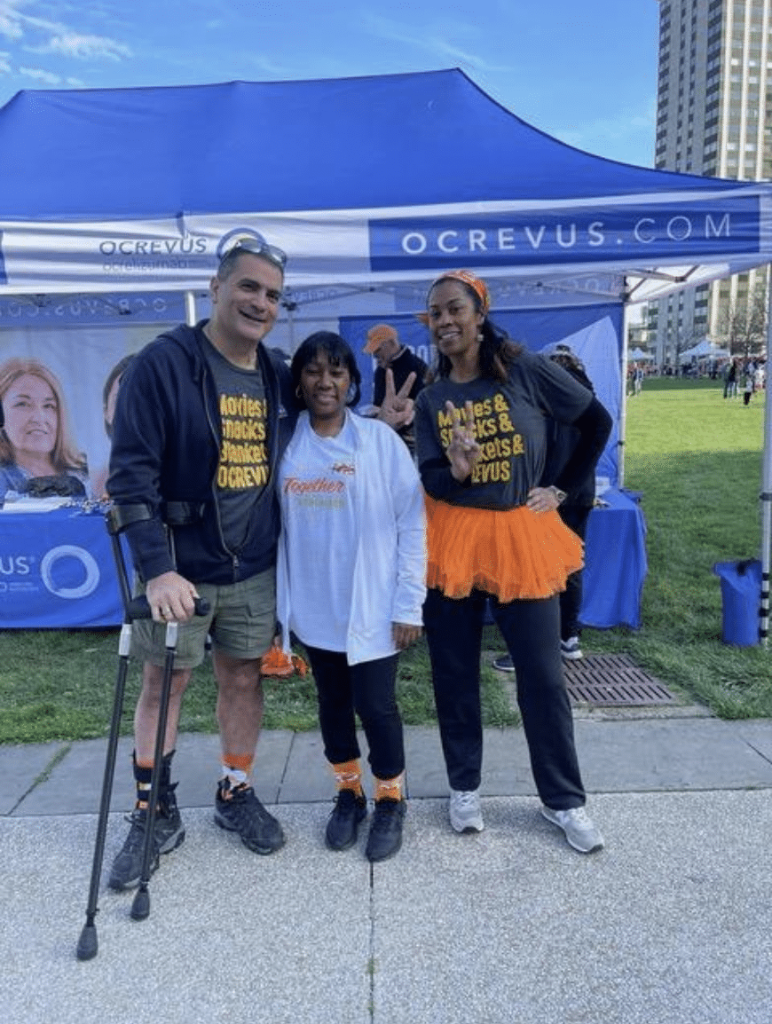 Three people smiling and posing at an outdoor event with a blue OCREVUS booth in the background. One person is using crutches, and the other two are wearing matching black shirts and orange accessories. Tall buildings and other event tents are visible behind them.