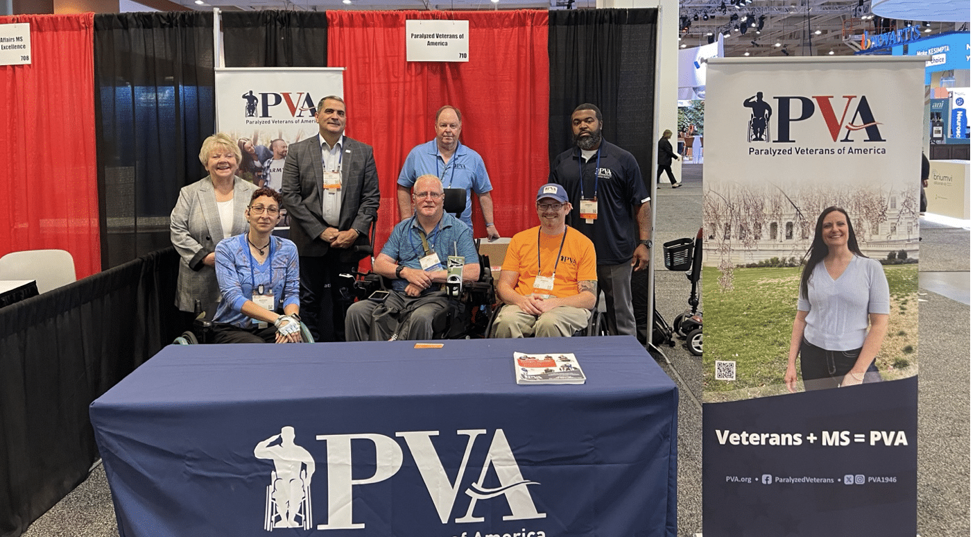 A group of eight people, some sitting and some standing, pose in front of a booth with "Paralyzed Veterans of America" signage and promotional materials. A table in the center has a blue cloth with the PVA logo. One side display features a woman and reads "Veterans + MS = PVA".