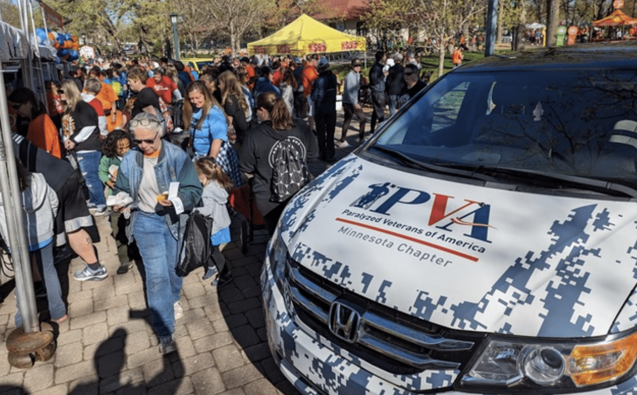 Outdoor community event with many people gathered around. In the foreground, a white van with "Paralyzed Veterans of America, Minnesota Chapter" written on it. Several people are engaging in activities and socializing under clear, sunny weather.
