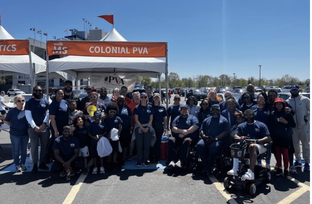 A large group of people, some in wheelchairs, pose together under and around a tent with the sign "COLONIAL PVA" at an outdoor event. They are wearing matching blue shirts and are smiling at the camera, with clear skies visible in the background.