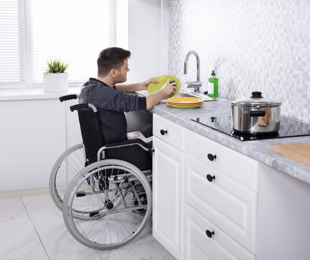 A person in a wheelchair is washing dishes at a kitchen sink. The kitchen has white cabinets, a marble countertop, and a tiled backsplash. There is a potted plant by the window, and a pot and cutting board on the counter.