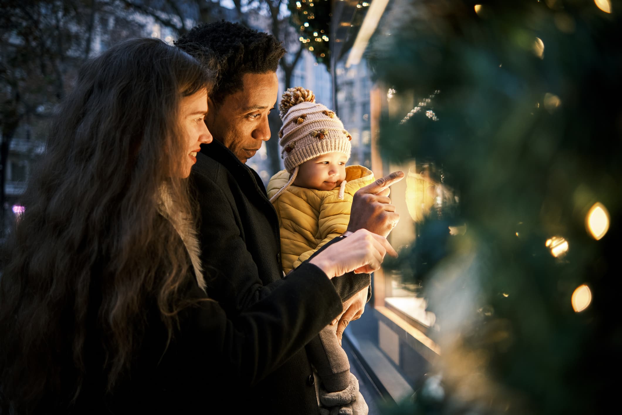 ImA family of three, consisting of a woman, a man, and a small child dressed in a yellow jacket and a knit hat, stands in front of a holiday-decorated store window, looking at the display with interest. The scene is illuminated with festive lights.