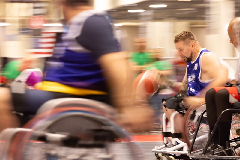 A man in a blue jersey intensely dribbles a basketball while maneuvering a wheelchair on a court. Motion blur highlights the fast-paced action as other players in wheelchairs race by. An American flag is visible in the background.