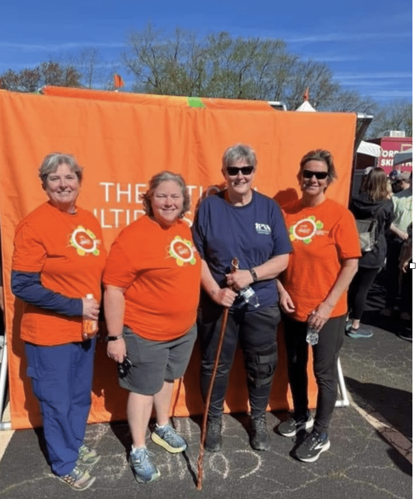 Four people stand and smile in front of an orange backdrop at an outdoor event. Three individuals wear orange shirts and one wears a navy-blue shirt while holding a walking stick. They appear to be participating in or supporting a walking event.