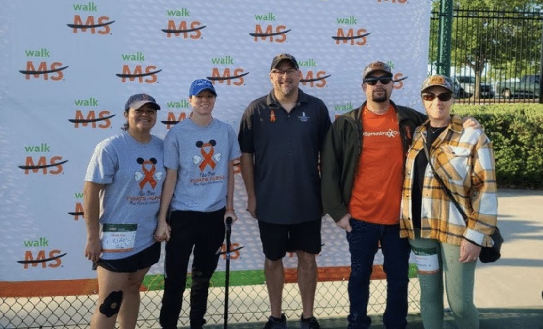Five people posing in front of a "Walk MS" backdrop. Two are wearing gray shirts with red ribbon designs, one in orange, and two in plaid. Three are standing, while two are leaning on canes. All appear to be participants in an MS awareness event.