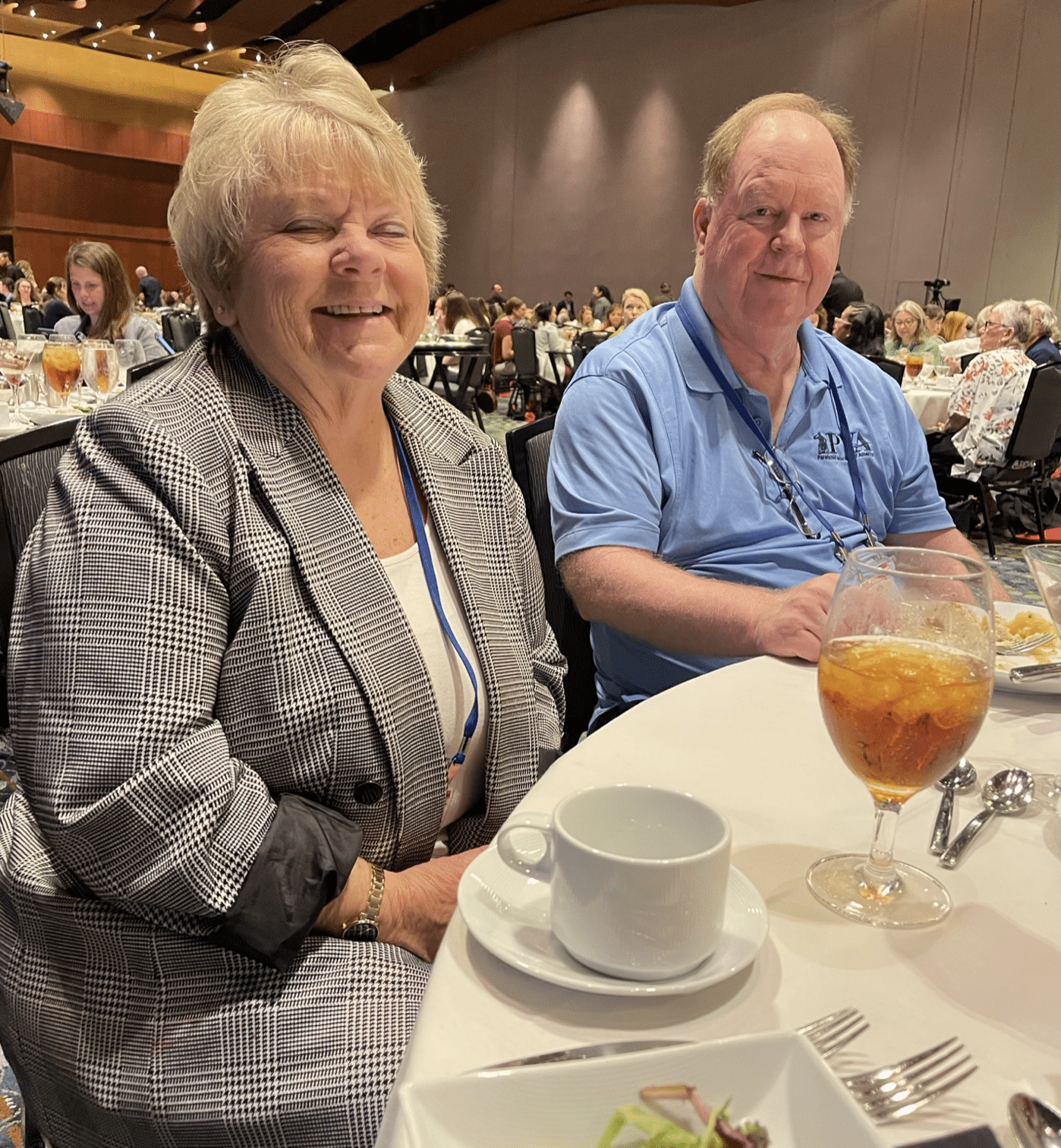 Two older adults, a woman and a man, are seated at a round table during a large gathering or conference. The woman is wearing a plaid jacket and has a name tag, while the man is in a light blue polo shirt. The table has a salad, iced tea, and a cup.
